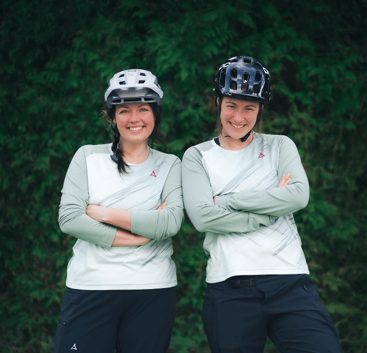 Zwei gleichgekleidete Frauen mit Fahrradhelmen lachen zusammenlehnend in die Kamera.
