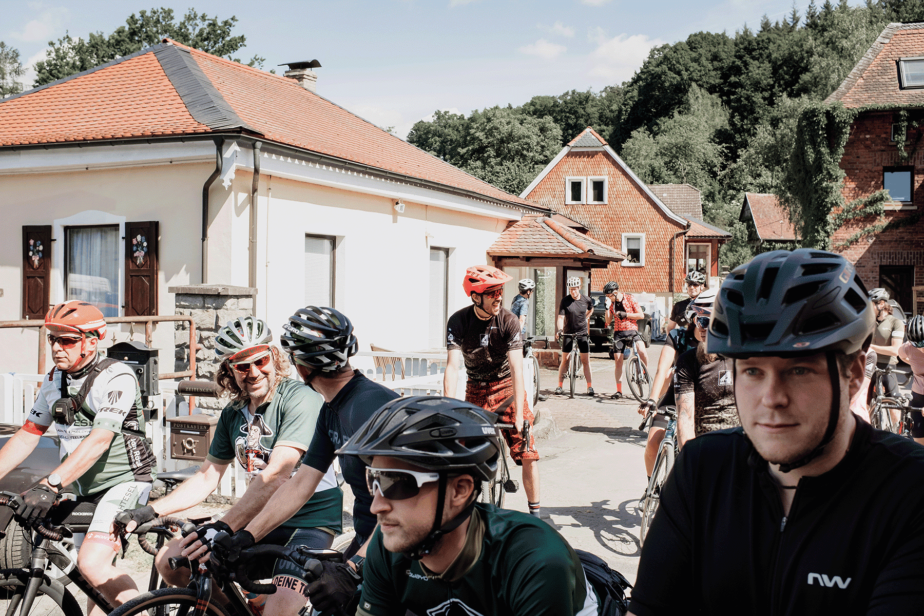 Eine Gruppe von Radfahrern mti Helmen steht in einer dörflichen Umgebung auf der Straße zwischen Häusern.